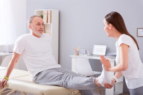 Real professional. Smiling male patient and a young female physiotherapist stretching patient foot being in a medical office