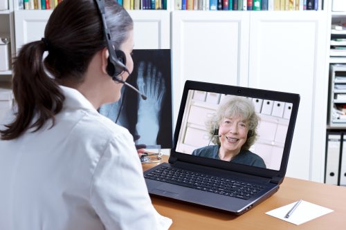 Female doctor in her surgery office with headset in front of her laptop, an x-ray of a foot in hand, talking with a senior patient, telemedicine concept